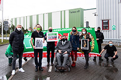 Kilkunastoosobowa grupa stoi w kurtkach i płaszczach na dworze na terenie firmy Armacell Poland w Środzie Śląskiej. Kilka osób trzyma flagę KKN. W tle budynki fabryczne.